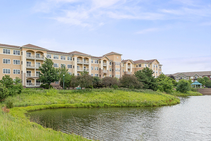 A pond in the foreground with a grass-covered embankment lined by trees backdropped by a four-story building with lots of windows and balconies. A gazebo with green roof sits in the distance next to the pond.
