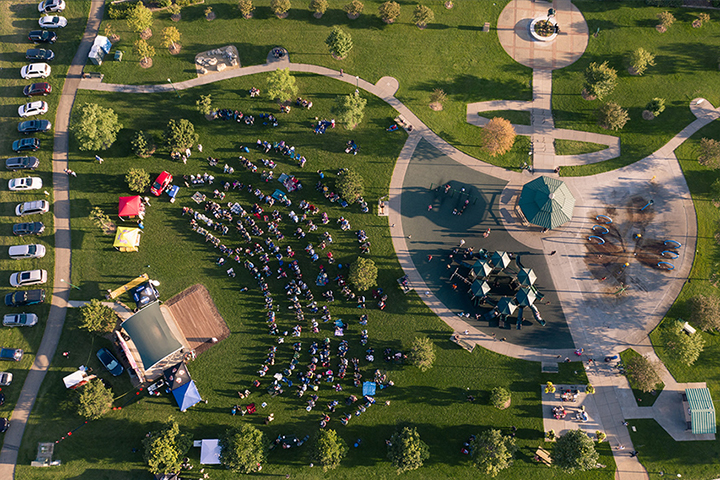 Aerial photo of Kelley Park during an outdoor music festival. Rows of people sit in chairs on the green grass in a semi circle facing the performance stage. The paved sidewalks create a circular design. The late afternoon sun casts long shadows across the park.
