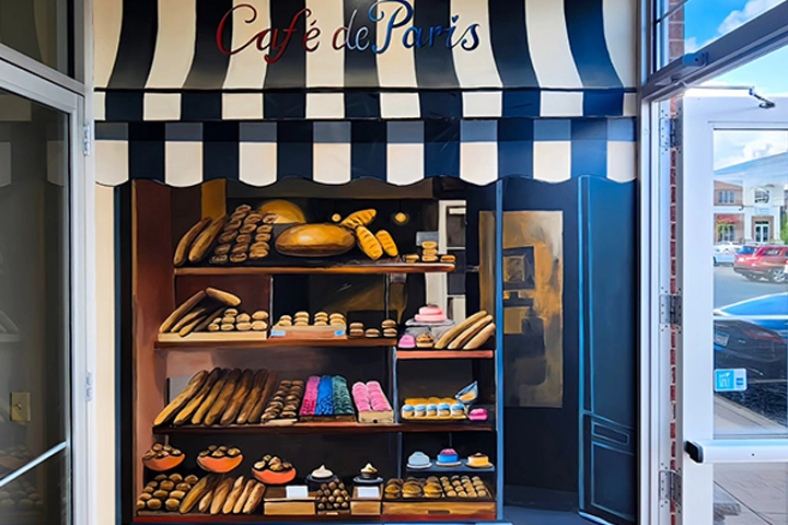 A mural on the entryway wall of a bakery, painted to look like the colorful display window of a french bakery, complete with black and white striped awning.