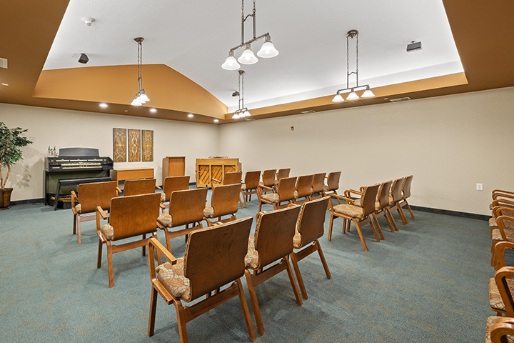A carpeted room with rows of upholstered armchairs facing the front of the room where an upright piano and an organ flank a wooden lecturn.