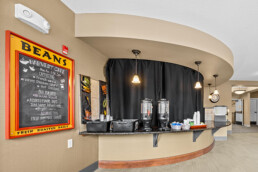 The curved countertop of a cafe holds two large coffee pots, cups and bussing trays. A chalkboard sign on the wall reads Beans Harvest Cafe with a list of menu items.