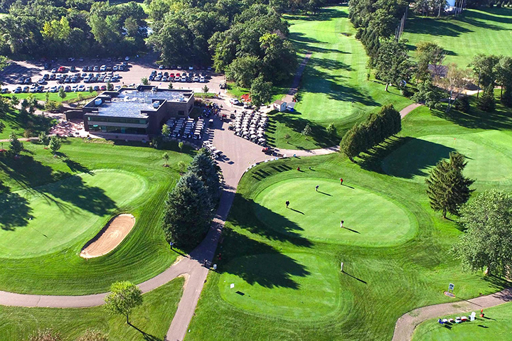 Aerial view of a golf club building surrounded by parking area, golf carts, a green, a tee box, a fairway, and mature trees.