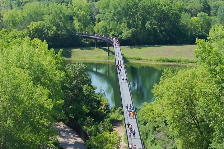 Aerial view of people walking on a raised walkway at the Minnestoa Zoo. The walkway crosses a pond and turns, disappearing into the tall trees.