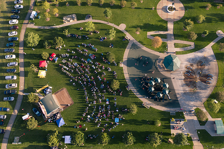 Aerial photo of Kelley Park during an outdoor music festival. Rows of people sit in chairs on the green grass in a semi circle facing the performance stage. The paved sidewalks create a circular design. The late afternoon sun casts long shadows across the park.