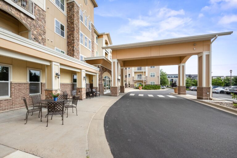 Exterior photo of carport over building main entrance with outdoor seating and table; blacktop driveway with striped crosswalk; four-story apartment building