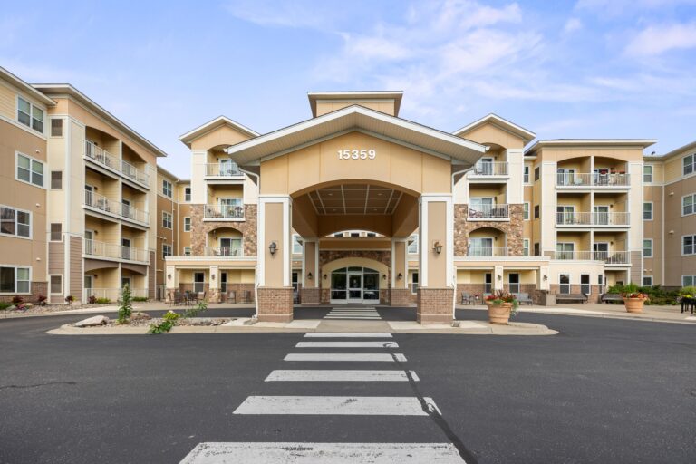 Exterior photo of carport over building main entrance; blacktop driveway with striped crosswalk; four-story apartment building with private balconies.