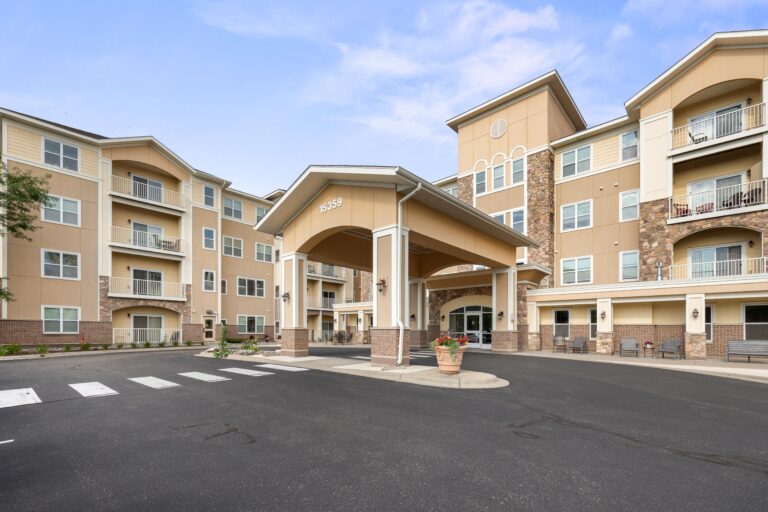 Exterior photo of carport over building main entrance; blacktop driveway with striped crosswalk; four-story apartment building with private balconies.