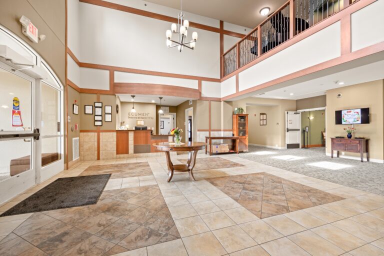 Entry way with large two-story atrium, chandelier, cream and brown tiled floor; concierge desk with Ecumen Seasons at Apple Valley logo on wall;