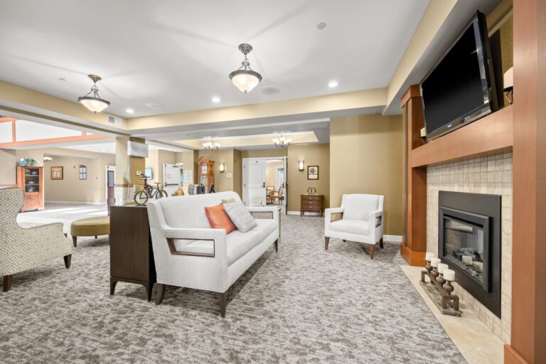 White sofa and matching lounge chair face modern wood and tile fireplace hearth with gas fireplace; additional lounge chairs and credenza sit behind the sofa; space is open and connects to hallways, entryways and meeting rooms.