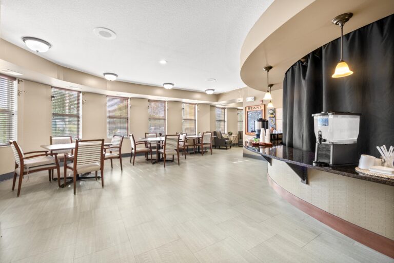 The curved countertop of a cafe holds a large ice water dispenser, cups, and straws; adjacent to a seating area with multiple four-person tables with upholstered chairs.