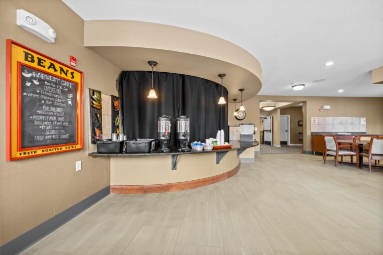 The curved countertop of a cafe holds two large coffee pots, cups and bussing trays. A chalkboard sign on the wall reads Beans Harvest Cafe with a list of menu items; adjacent to a seating area with four-person tables and upholstered chairs..