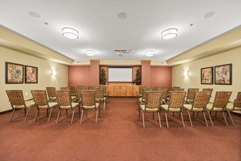 Large, carpeted room with rows of upholstered arm chairs and an ceiling-mounted projector facing a wall featuring a white screen and buffet built-in cabinet.