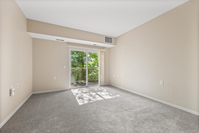 An unfurnished room with light-colored carpet, cream walls, and vertical blinds on a sliding glass door leading to a private balcony overlooking trees.