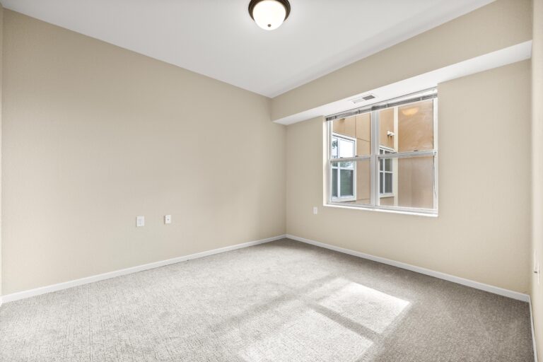An unfurnished bedroom with light-colored carpet, cream walls, and large window with blinds.