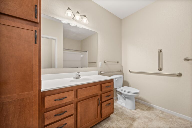 Large bathroom with wood cabinetry, grab bars, toilet and long vanity with sink, mirror, and drawers.