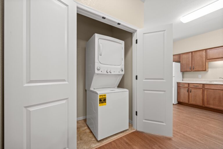 An unfurnished apartment with white in-unit stacked washer and dryer with dedicated laundry closet and double white doors; next to wood flooring and kitchen with white appliances and wood cabinetry.