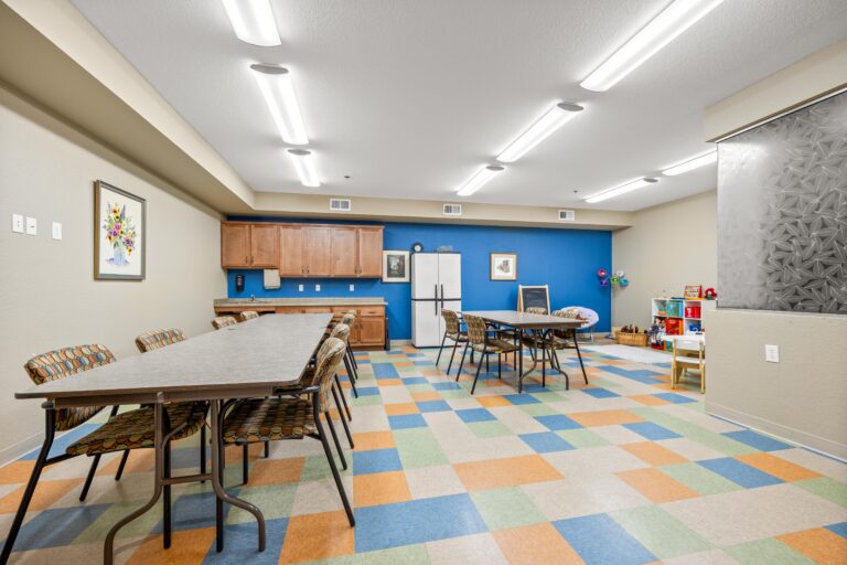 Colorful room with multi-color tile floor and two long task tables with upholstered chairs; sink, counter and cabinets; refrigerator; kids craft and play area.