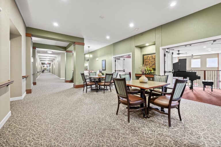 Large, carpeted hallway with sitting area, featuring sage green walls, two task tables and upholstered chairs; adjacent to double doors opening to another sitting area with black baby grand piano.