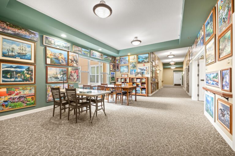 Large, carpeted sitting area with two task tables and chairs; book shelf full of puzzle boxes, and sage green walls covered floor-to-ceiling with completed, framed puzzles.