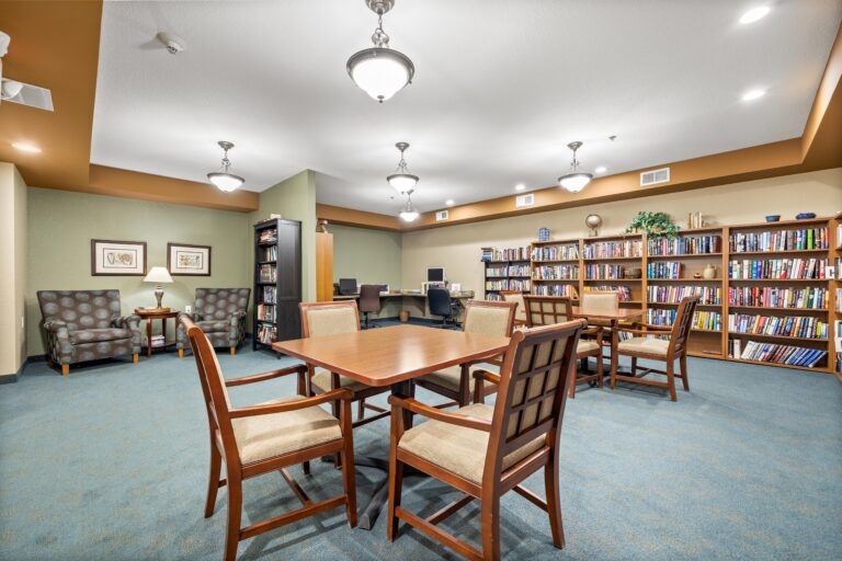 Bright room with blue-green carpeting, two task tables and upholstered chairs, lounge chairs, a corner computer center, and a wall of full book shelves.
