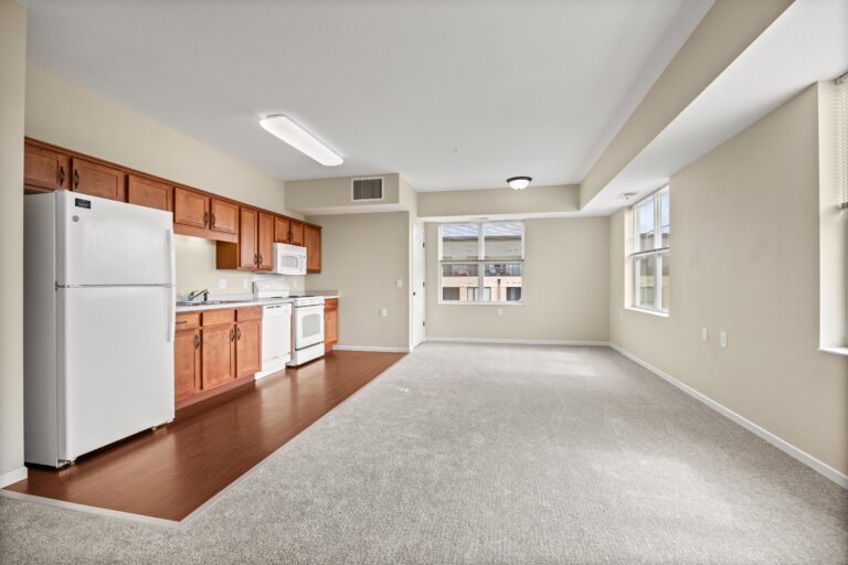 The full kitchen space with wood flooring, white refrigerator, range, microwave, and dishwasher appliances and stained wood cabinetry; adjacent to light-colored carpeting in living area with large windows and blinds.