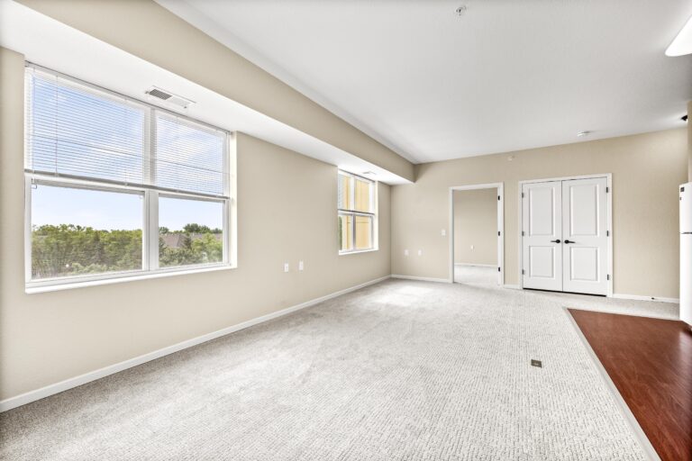 An unfurnished room with light-colored carpet, cream walls, large windows with blinds, doorway to a bedroom, white laundry closet doors, and the edge of kitchen flooring adjacent to living room area.
