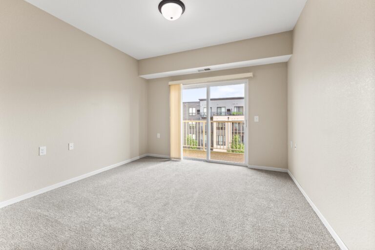 An unfurnished room with light-colored carpet, cream walls, and vertical blinds on a sliding glass door leading to a private balcony.
