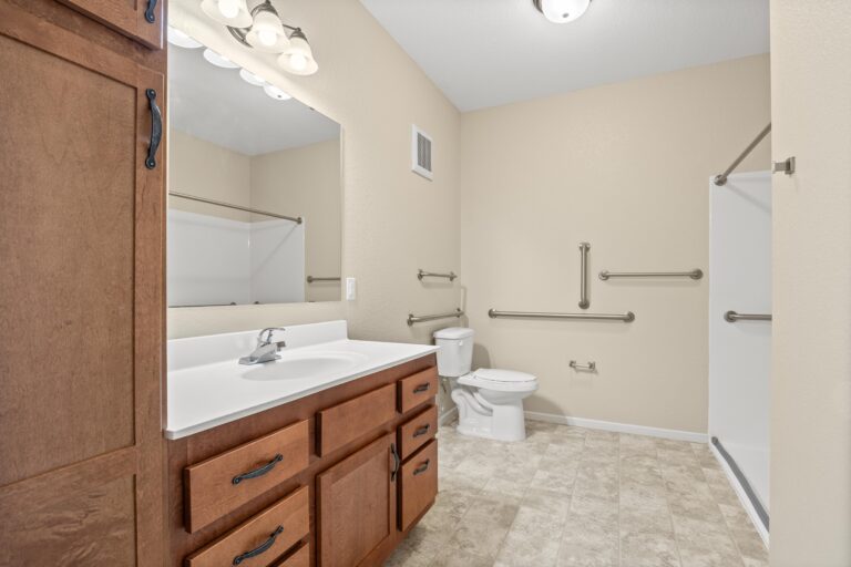 Large bathroom with wood cabinetry, grab bars, walk-in shower, toilet and long vanity with sink, mirror, and drawers..