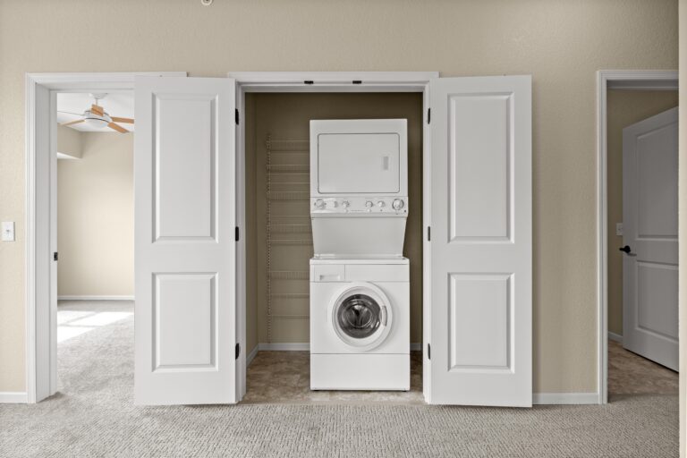 An unfurnished apartment with light-colored carpet floor, white in-unit stacked washer and dryer with dedicated laundry closet and double white doors; doorway to bedroom with sunlight and ceiling fan; doorway to bathroom.
