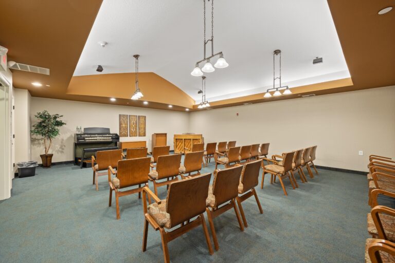 A carpeted room with rows of upholstered armchairs facing the front of the room where an upright piano and an organ flank a wooden lecturn.