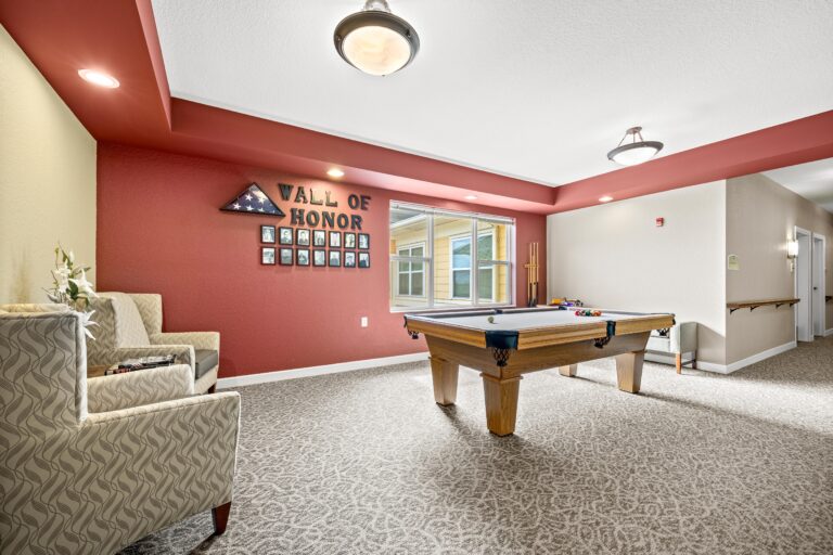 A billiards table in a well-lit area next to upholsterd lounge chairs and a crimson wall featuring a Wall of Honor with a folded and framed American flag hung next to photographs of late Veteran residents.