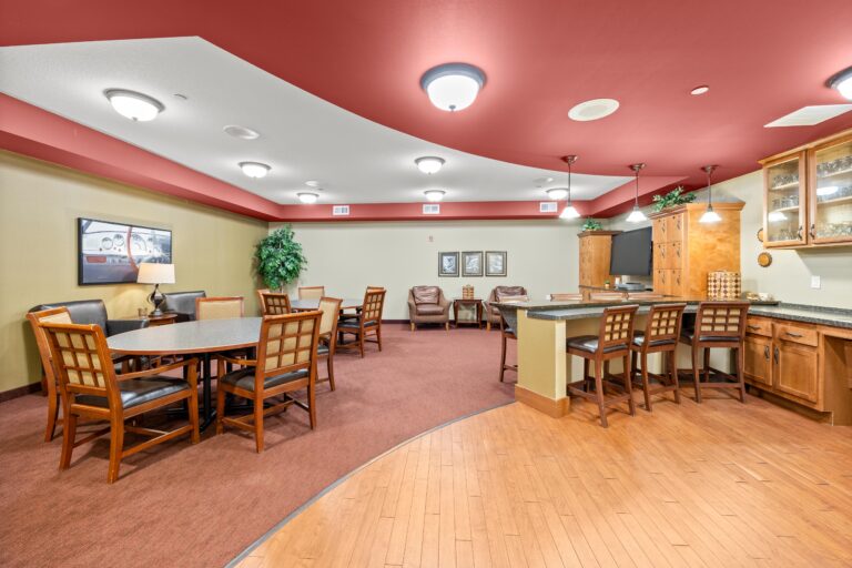 Carpeted lounge area with two dining tables and upholstered seating, lounge seating, television, and personal lockers; bar with barstools; kitchen with small refrigerator, sink, glass-door cabinets and pendant lighting.