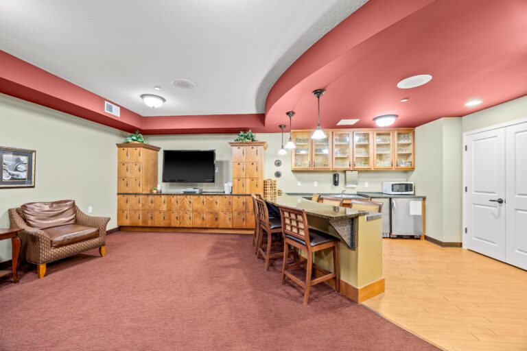 Carpeted lounge area with upholstered seating, television, and personal lockers; bar with barstools; kitchen with small refrigerator, sink, glass-door cabinets and pendant lighting.