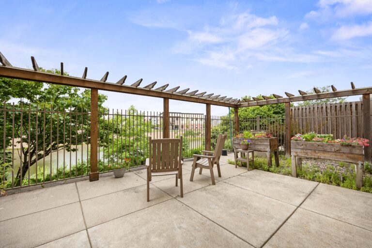 A secured outdoor patio with seating, raised garden beds, and pergola surrounded by trees.
