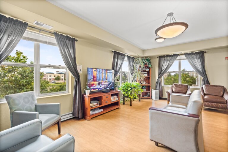 A bright room with many windows, upholstered lounge chairs, a television, and a games cabinet.