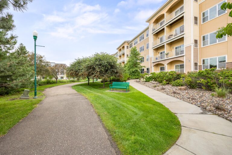 A paved, treelined walkway passes a park bench and connects to a sidewalk leading to a four-story apartment building.