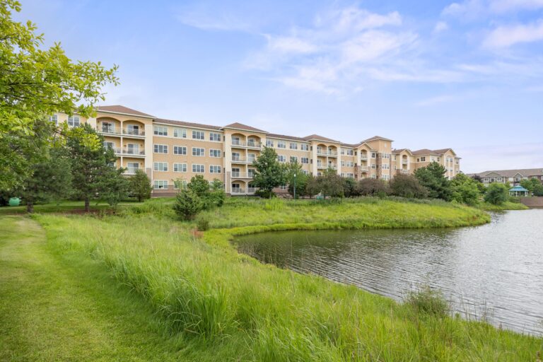 A pond in the foreground with a grass-covered embankment lined by trees backdropped by a four-story building with lots of windows and balconies. A gazebo with green roof sits in the distance next to the pond.