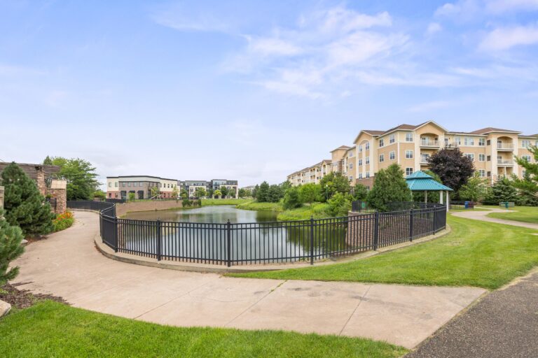 Next to a four-story apartment building, a paved walkway stretches from a park area and wraps around the end of a narrow pond and continues toward other buildings on the pond's edge.