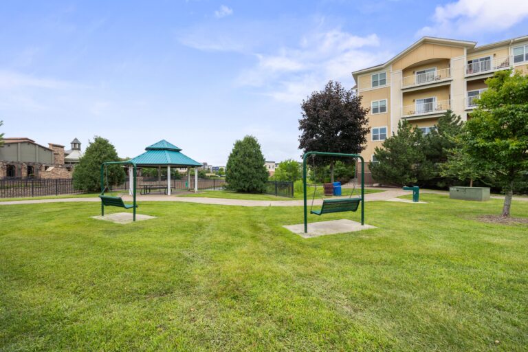 Next to a four-story apartment building is a park area with green grass, two swinging benches, a paved walkway, and a sheltered picnic table..