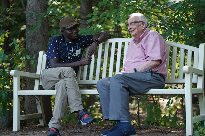 A younger male team member sits on a white bench outside next to an older gentleman as they engage in discussion.