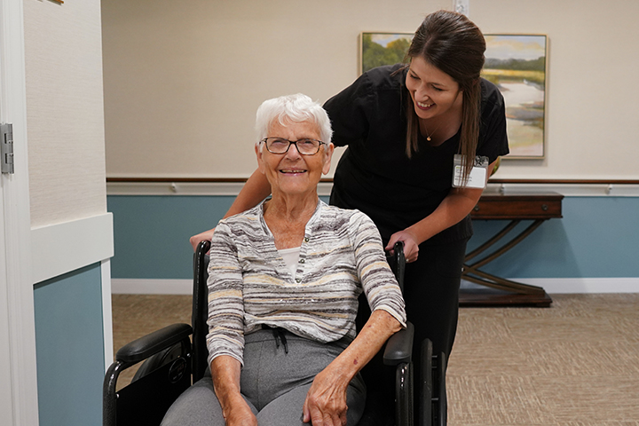 A young female caregiver wearing scrubs and an name tag, smiles as she pushes a smiling older women in a wheelchair down a hallway..