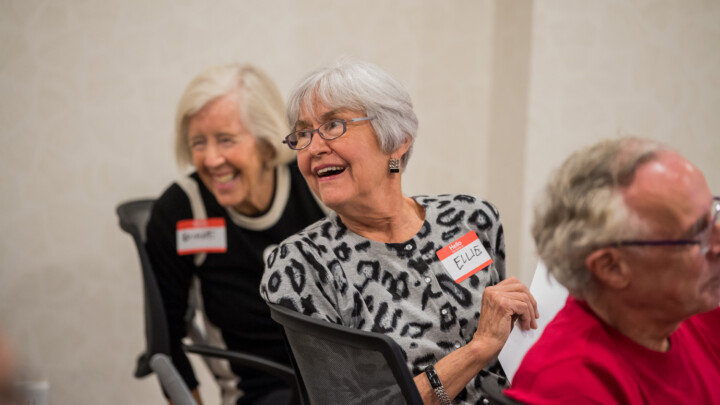 3 senior volunteers in chairs looking behind them smiling and wearing name tags