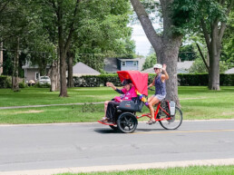 Marcia Kull driving a trishaw bike (a three-wheeled bike that can carry passengers) waving with 2 smiling a waving residents of Ecumen Pathstone.