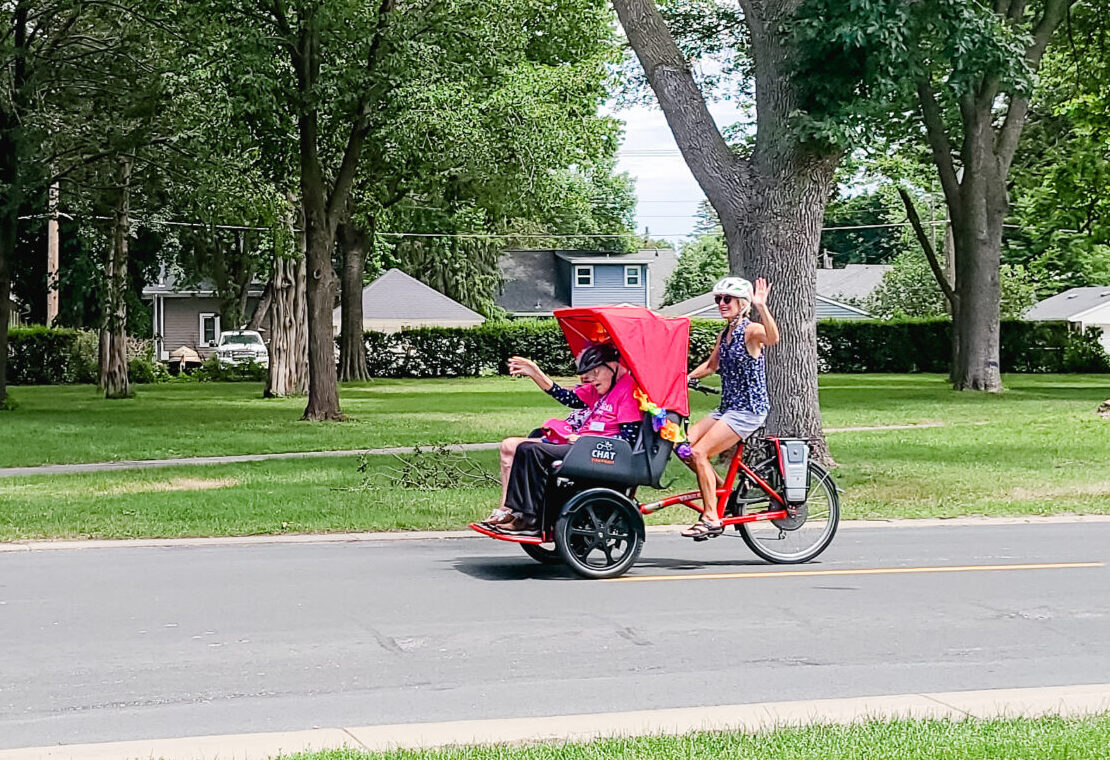 Marcia Kull driving a trishaw bike (a three-wheeled bike that can carry passengers) waving with 2 smiling a waving residents of Ecumen Pathstone.