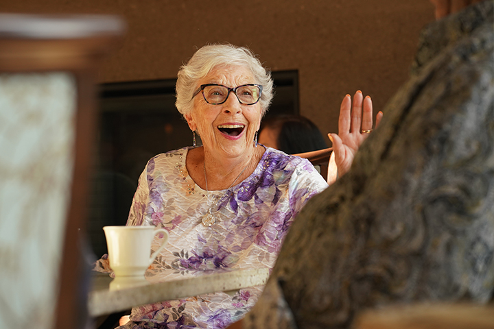 An older woman sitting at a dining table and wearing a floral patterned blouse waves and smiles at someone at a nearby table.