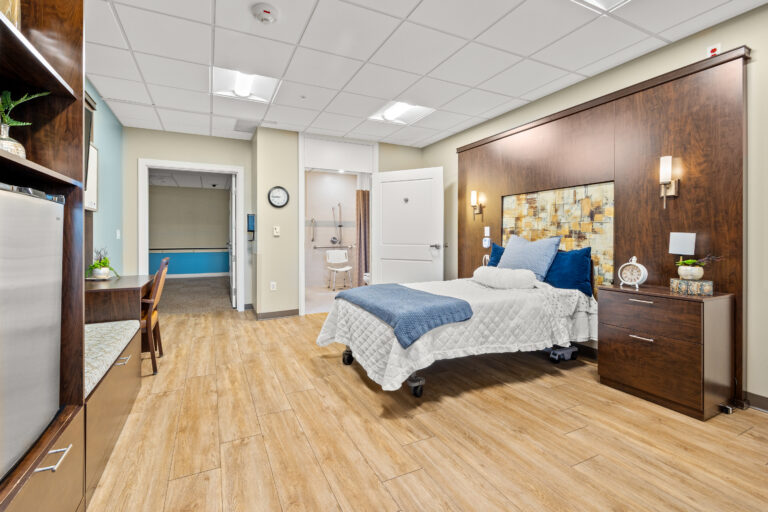 Bright, modern Memory Care suite at Chateau Waters featuring a single bed with white linens and blue accents, wooden floors, a bathroom door open, and a desk on the left.