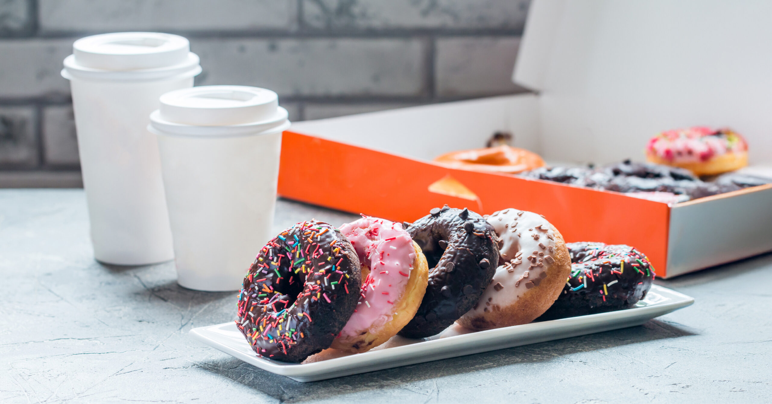 A plate of colorful donuts with sprinkles sits on a table next to two takeaway coffee cups. An open box of donuts is visible in the background.