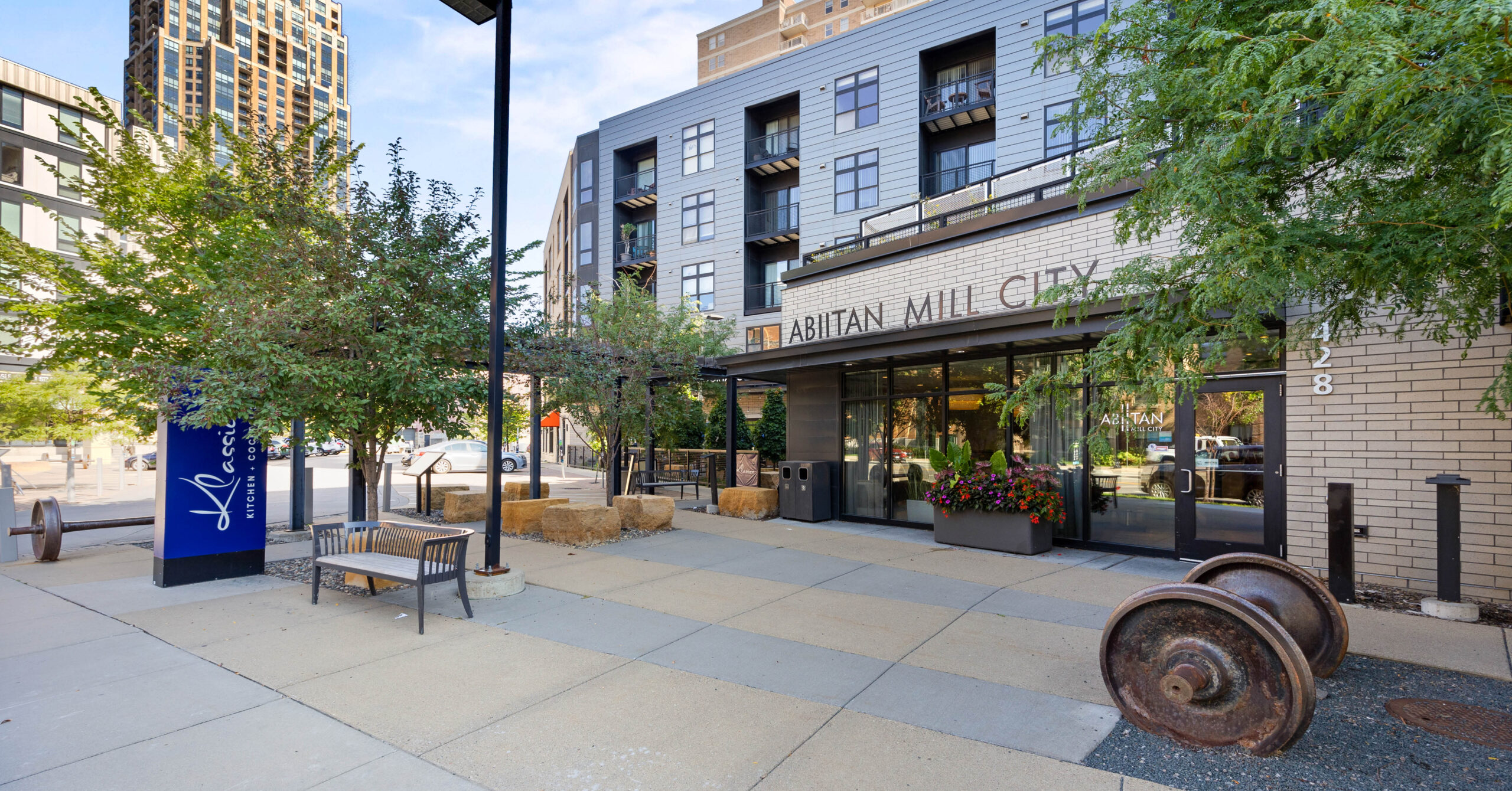 Modern urban setting in downtown Minneapolis outside a brick building labeled "ABIITAN MILL CITY," with trees, benches, and industrial decor creating a welcoming ambiance.
