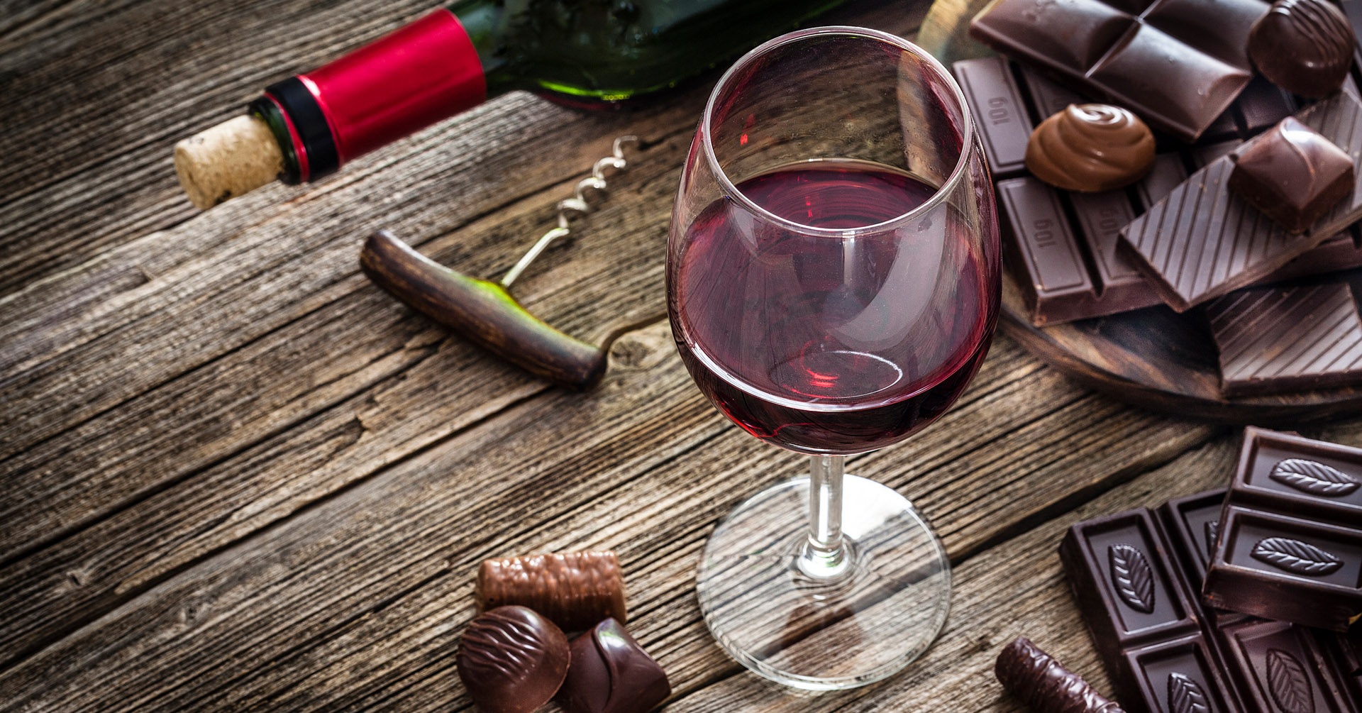 A glass of red wine is surrounded by assorted chocolates on a rustic wooden table. A corkscrew and wine bottle lie nearby, creating a cozy, indulgent scene.
