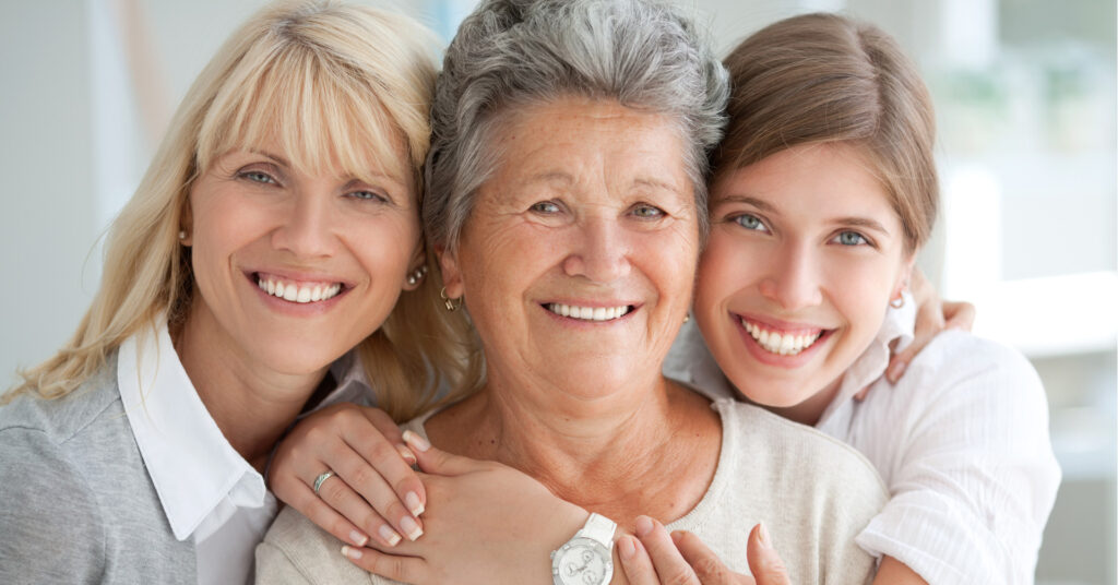 Three women of different generations smile warmly at the camera. The older woman is in the center, embraced by two younger women on each side, conveying love and togetherness.